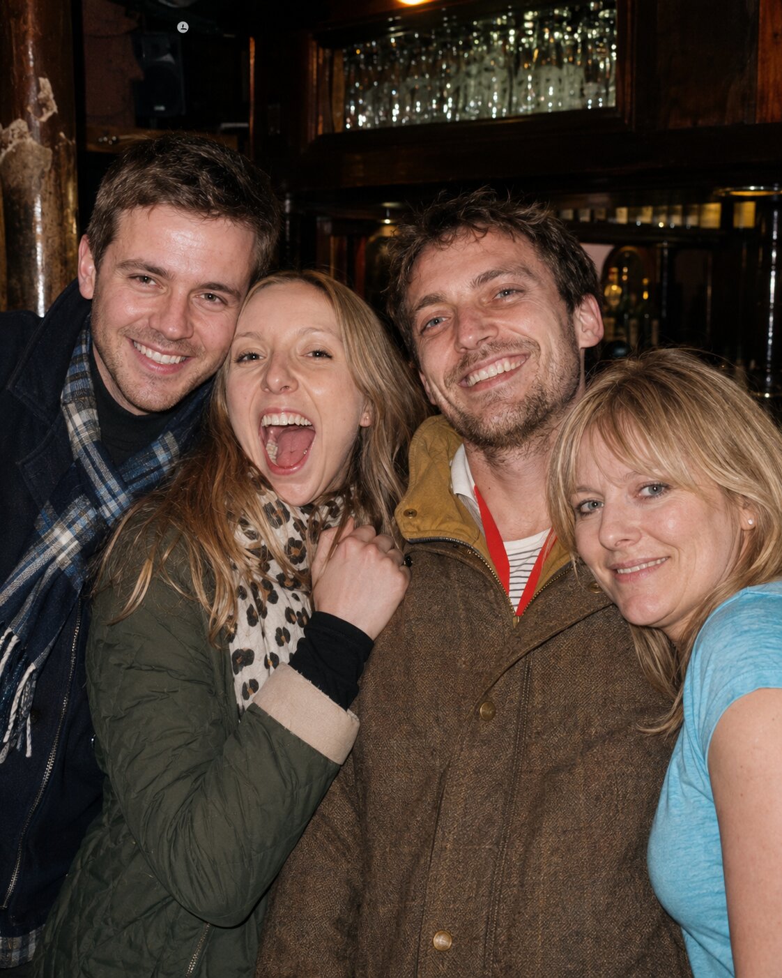 A younger Élise and Mateo laughing with two friends, crowded together in a warm wood-panelled bar the night they met